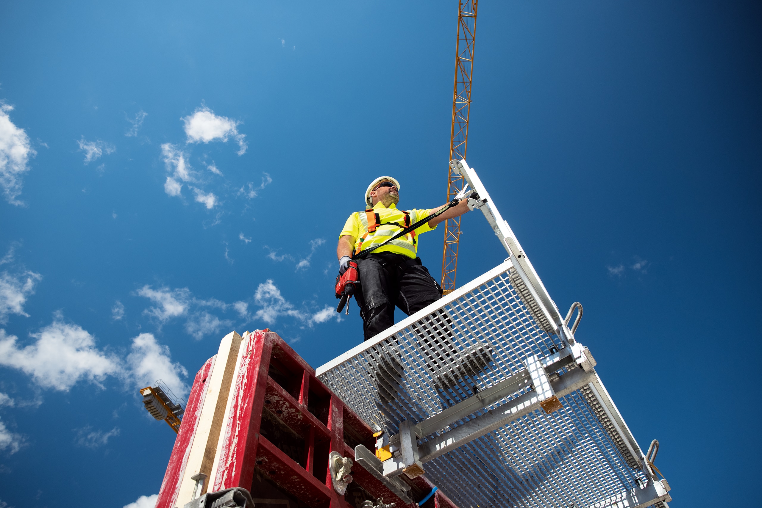 Worker standing on a platform with a drill in his hand that is fixed to a pole with a tool tether