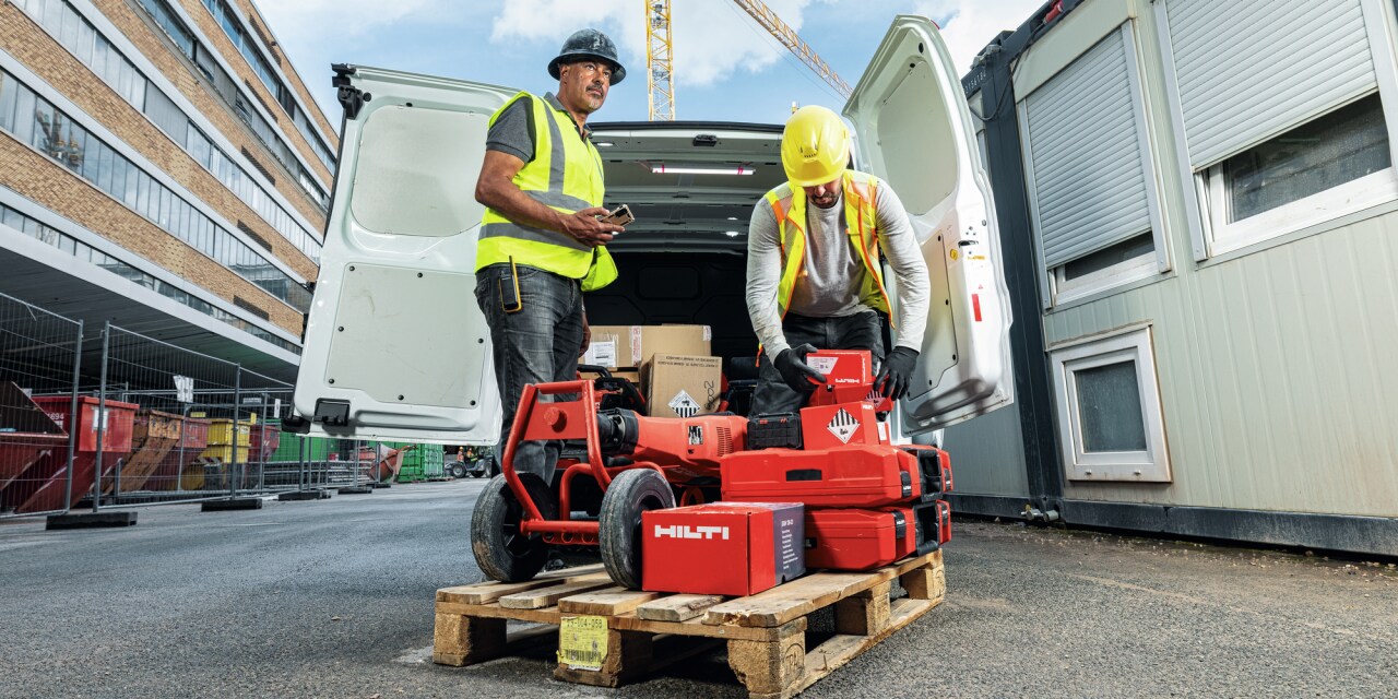 Two men moving red Hilti tool boxes from a wooden pallet into a white van. 