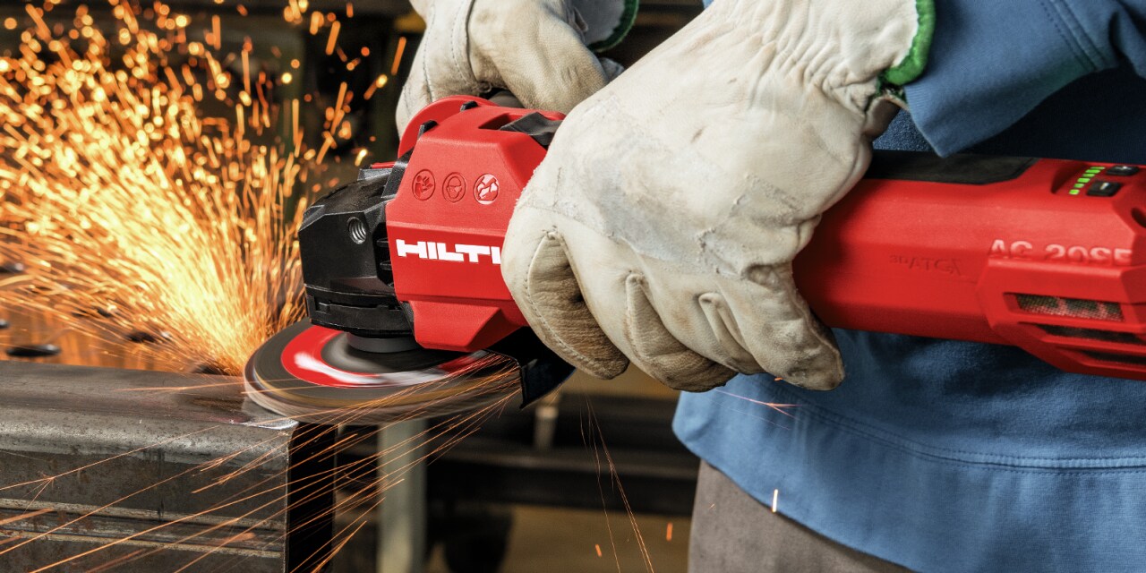 Man in safety gloves using red Hilti angle grinder, cutting metal rebar. Sparks are in the air. 