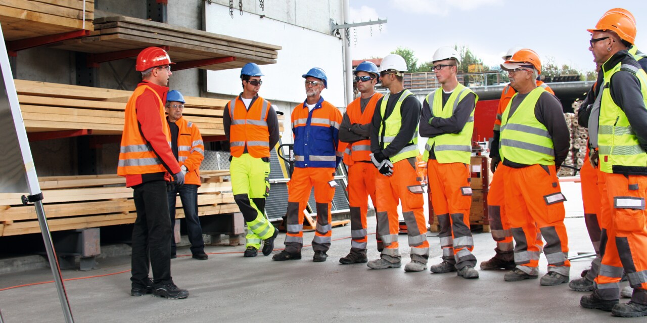 Group of construction workers in high visibility vests having a briefing inside of a warehouse.