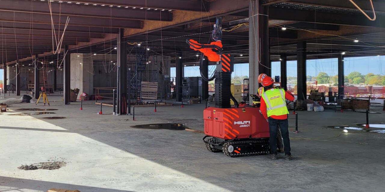 Red Hilti Jailbot is working on construction site. One construction worker in yellow high visibility vest is standing beside.